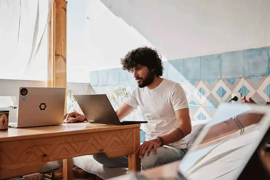 A person with curly hair works intently on a laptop at a wooden table, surrounded by bright decor and natural light in Sundesk cowork in Taghazout.