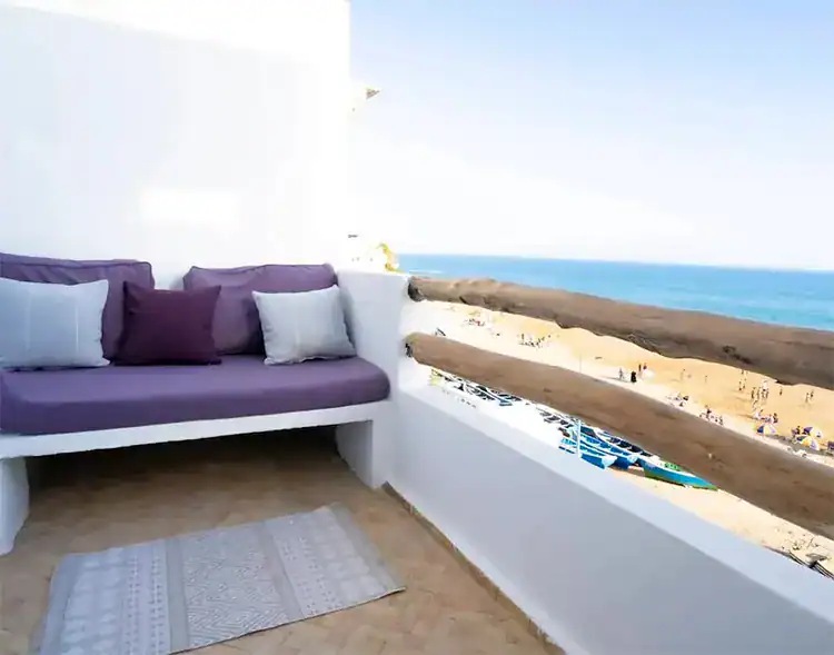Cozy balcony with a purple couch and cushions overlooking a sandy beach and blue ocean under a clear sky at Sundesk Taghazout.