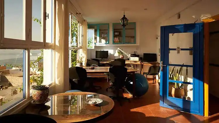 Bright office space with large windows, modern furniture, computers, potted plants, and colorful blue door, showcasing a scenic view at Sundesk in Taghazout.
