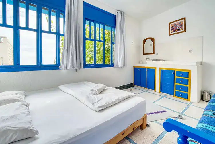 Bright room featuring a double bed, a blue sofa, and colorful tile accents, with a warm wooden door and decorative lighting at Sundesk in Taghazout.