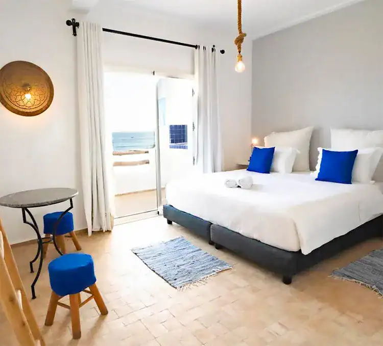 Bright, modern bedroom with a double bed, blue accents, minimalist decor, a table, and a balcony view of the ocean in Sundesk Taghazout.