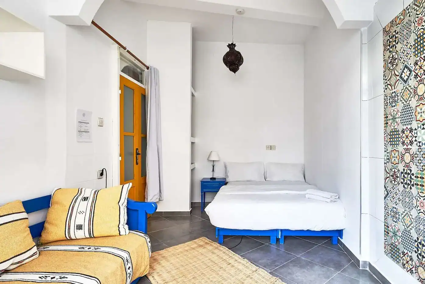 A cozy room featuring a double bed, blue furniture, elegant decor, and a vibrant tiled wall; sunlight streams through an orange door in Sundesk Taghazout. 