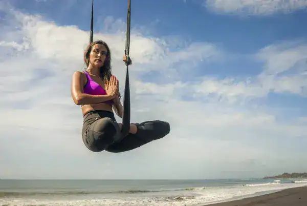 A woman practices aerial yoga on a beach, suspended in a pose with waves and a cloudy sky in the background in Taghazout.
