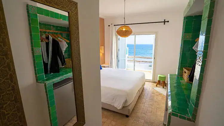 Bright bedroom with a view of the ocean, featuring green tile accents, a cozy bed, and a stylish mirror reflecting hanging clothes at Sundesk in Taghazout.