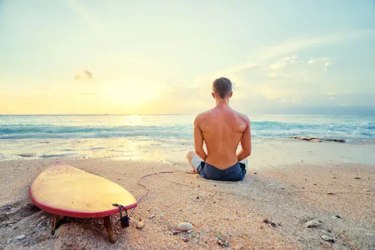 A man sitting and meditating by the beach with his surfboard in Taghazout.