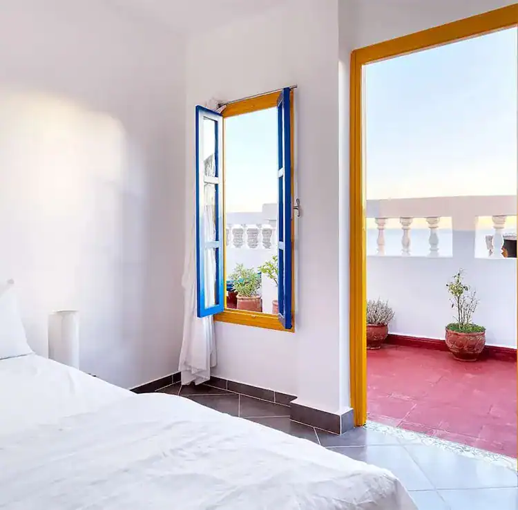 Bright bedroom with a white bed, blue window frames, and a doorway leading to a sunny terrace with plants at Sundesk in Taghazout.